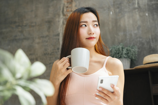 年轻的亚洲女性早餐喝咖啡，手持智能手机。她从右边看。