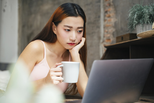 年轻的亚洲女人看着笔记本电脑，手里拿着一杯咖啡。