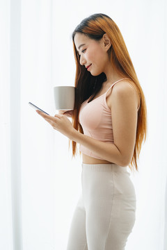 侧视图-年轻的亚洲女性拿着一杯咖啡，在白色窗帘前使用智能手机。