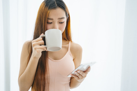 美丽的亚洲年轻女子，手拿咖啡，在白色背景下使用智能手机。