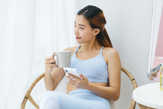 美丽的运动型亚洲女性，穿着运动胸罩，手持咖啡杯和智能手机。