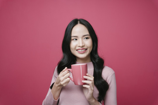 粉红色背景下，年轻的亚洲女性手持咖啡杯的肖像。