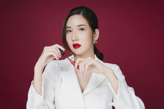 身着白色西装夹克、在猩红色背景上涂上红色唇膏的美丽年轻亚洲女性肖像。