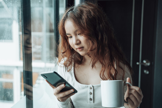 亚洲女性早上在窗户边用智能手机喝热咖啡。