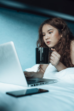 年轻的亚洲女性在宿舍的卧室里使用笔记本电脑和热咖啡。