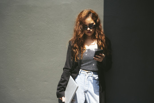 戴着墨镜和黑色夹克的年轻亚洲女性使用智能手机的时尚肖像。