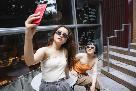 两位年轻的亚洲女性戴着太阳镜在户外咖啡馆用智能手机自拍的照片。