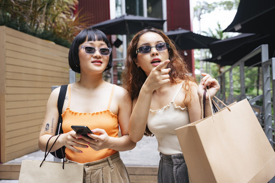 两位戴着太阳镜的年轻亚洲女性在街头购物中心寻找销售店的照片。
