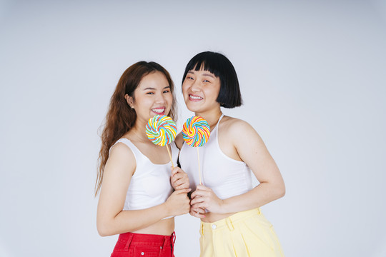 两位身穿白色背心、手持彩虹糖果棒棒糖的亚洲年轻女性的肖像，背景为白色。