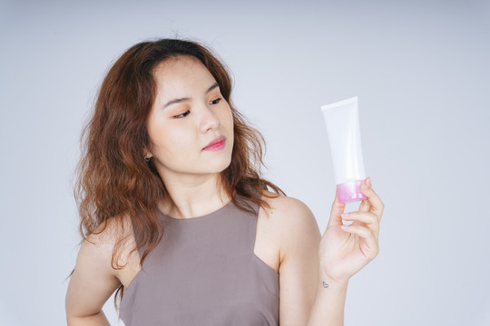 白色背景下，年轻的亚洲卷发女性手持护肤霜乳液瓶的肖像。