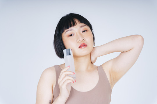 亚洲年轻短发女性的特写镜头，手持乳液或香水瓶，背景为白色。