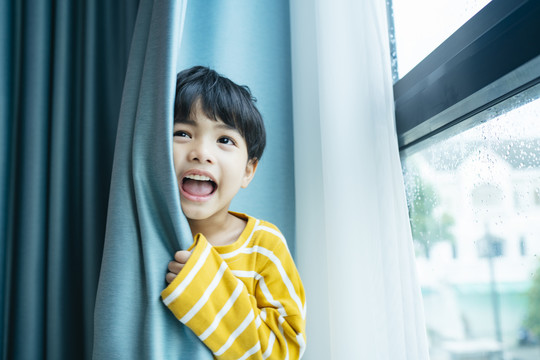 在靠近窗户的蓝色窗帘后面，一个穿着白色毛衣的快乐有趣的男孩的肖像。