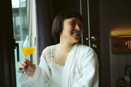 身穿白色浴袍的亚洲短发女子在酒店房间享用橙汁早餐。