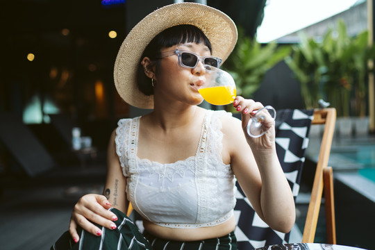 一位戴着太阳镜和帽子在酒店喝橙汁的亚洲年轻女子。