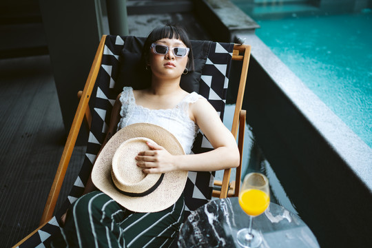 年轻的亚洲女人戴着墨镜和帽子躺在酒店游泳池附近的沙滩椅上。