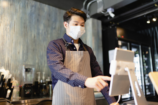 戴着面具的亚洲咖啡师在笔记本电脑上操作数字菜单。2019冠状病毒疾病概念后重新开放业务。