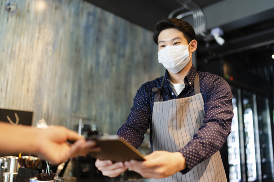 一名戴着面具的亚洲咖啡师在咖啡馆向顾客点菜。2019冠状病毒疾病概念后重新开放业务。