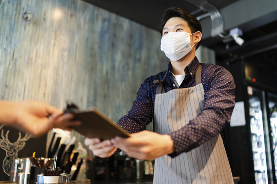 一名戴着面具的亚洲咖啡师在咖啡馆向顾客点菜。2019冠状病毒疾病概念后重新开放业务。