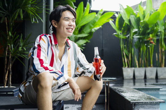 兴高采烈的亚洲年轻人坐在家里的游泳池旁，享受着一瓶美酒。