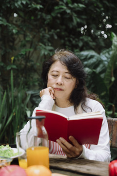 一位亚洲老妇人在花园里的野餐桌旁看书。