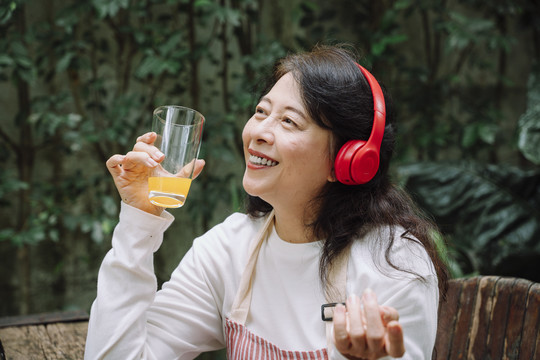 亚洲资深女性戴着无线耳机，手持橙汁听音乐。