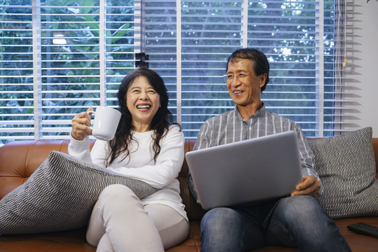 快乐的亚洲老年夫妇坐在沙发上，用数字笔记本电脑浏览网站。