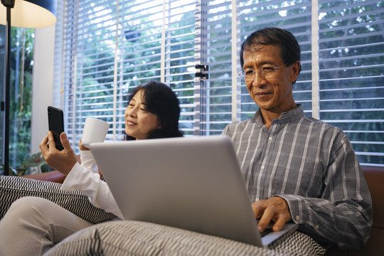 快乐的亚洲老年夫妇坐在沙发上，使用智能手机和笔记本电脑。