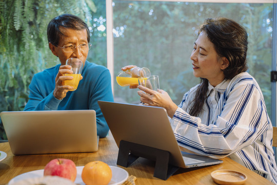 亚洲的一对老年夫妇在使用笔记本电脑时喜欢喝orage果汁。