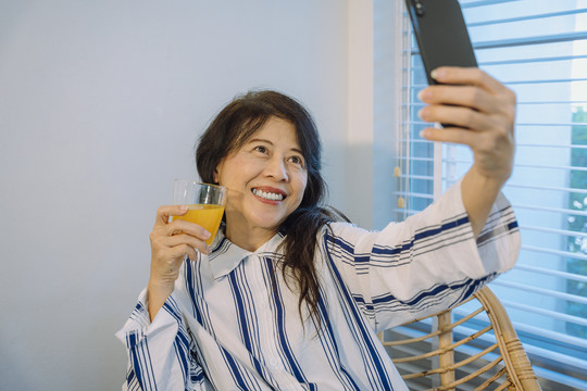 拿着橙汁用智能手机自拍的亚洲资深女性。