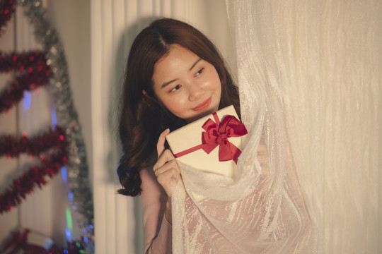 美丽的亚洲年轻女子黑发在白色窗帘后拿着圣诞礼品盒。