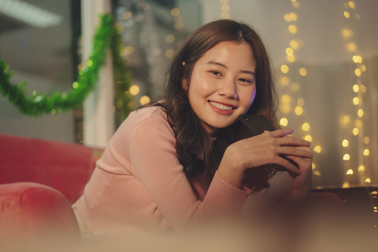在圣诞派对上，一位年轻的亚洲美女黑发坐在沙发上，手里拿着智能手机。