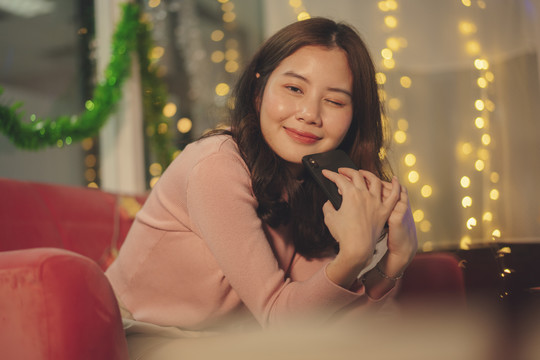 在圣诞派对上，一位年轻的亚洲美女黑发坐在沙发上，手里拿着智能手机。