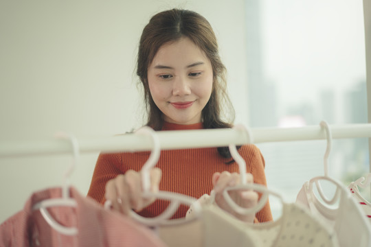 美丽的亚洲年轻女子黑发在家里从衣架上挑选衣服。