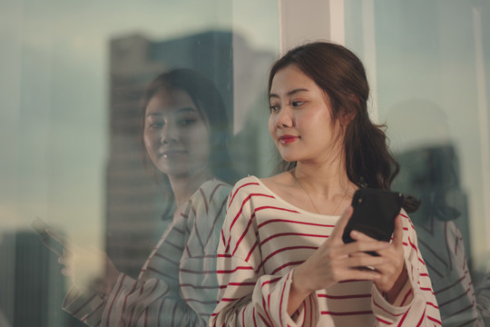 美丽的亚洲年轻女子穿着毛衣，在窗户附近用智能手机梳着黑发。