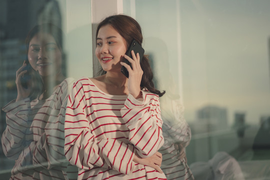 美丽的亚洲年轻女子穿着毛衣，在窗户附近用智能手机梳着黑发。