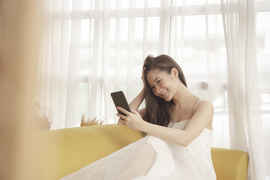 年轻的亚洲美女穿着白色衣服，在客厅的沙发上使用智能手机。