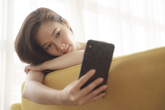 年轻的亚洲美女在家里用智能手机在沙发上自拍。