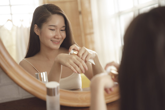 年轻的亚洲美女在镜子前的脸上涂抹护肤霜。