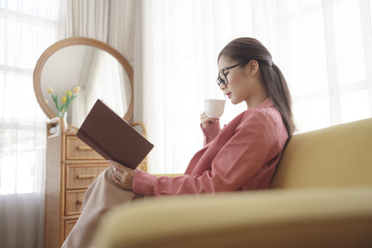 侧视图——年轻的亚洲美女穿着夹克，戴着眼镜，喝着咖啡，在沙发上看书。