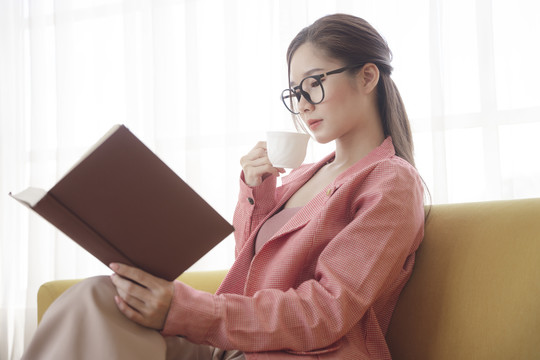 侧视图——年轻的亚洲美女穿着夹克，戴着眼镜，喝着咖啡，在沙发上看书。