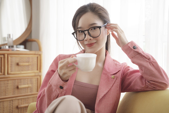 年轻的亚洲美女穿着粉色夹克，戴着眼镜，手里拿着一杯咖啡。