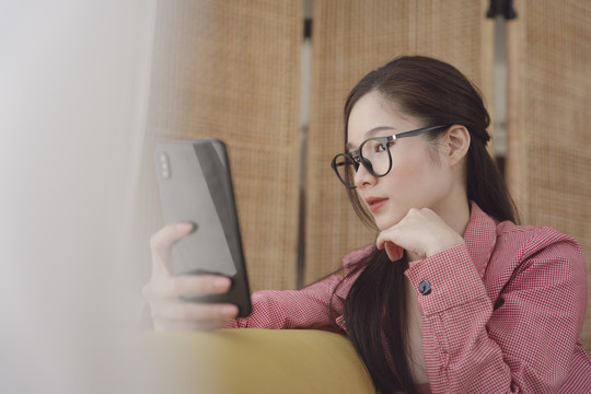 侧视图-年轻的亚洲美女戴着眼镜，通过智能手机观看视频或直播。