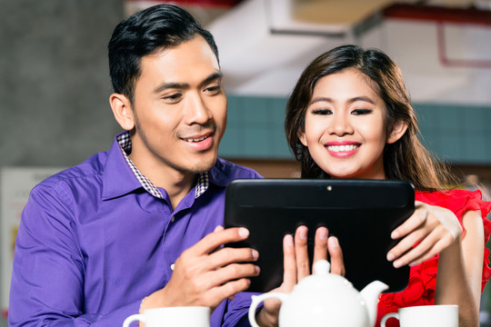 一对年轻快乐的印尼夫妇在一家时髦的咖啡店用平板电脑一起观看在线有趣的视频