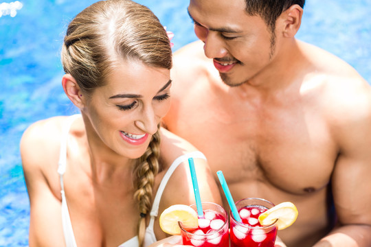 一对正在度假的亚裔男子和白人女子，在热带花园里，在酒店的游泳池里用饮料或鸡尾酒洗澡