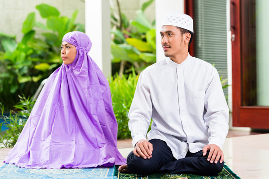一对亚洲穆斯林夫妇，一男一女，坐在热带花园前他们家的祈祷地毯上，在家里祈祷