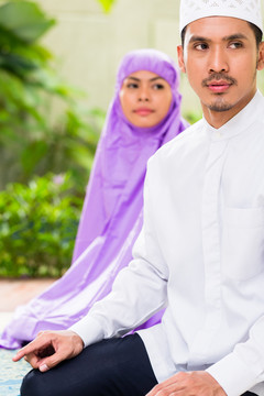一对亚洲穆斯林夫妇，一男一女，坐在热带花园前他们家的祈祷地毯上，在家里祈祷