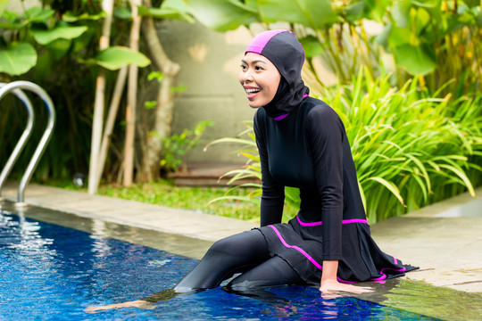 穿着布基尼清真泳衣的穆斯林妇女或女孩坐在热带花园的游泳池旁