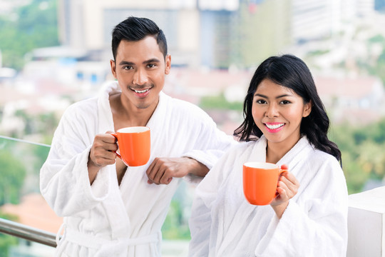 一对亚洲夫妇早上站在城市天际线前，穿着浴衣喝着咖啡