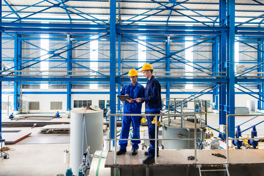 两名工人站在大型机器上，在大型金属车间或工厂检查工作