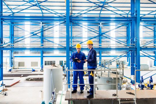 两名工人站在大型机器上，在大型金属车间或工厂检查工作